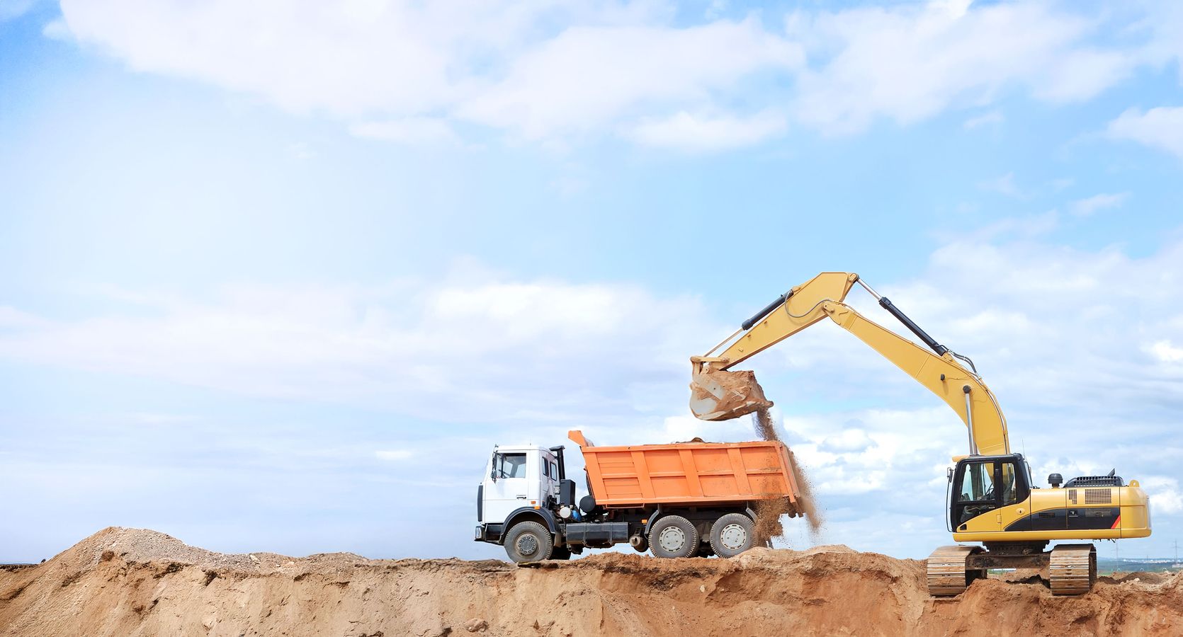 Bagger lädt Erde in einen orangefarbenen Muldenkipper auf einer Baustelle unter blauem Himmel